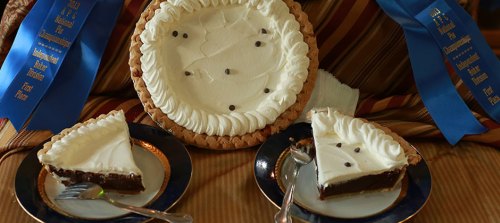 A chocolate cream pie with whipped cream and chocolate chips, accompanied by two slices on plates with silver forks.