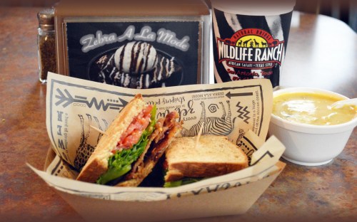 A sandwich with lettuce, tomato, and bacon, served with a cup of soup and a drink, on a patterned tray.