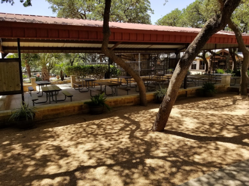 Outdoor seating area with tables under a shaded pavilion, surrounded by trees and gravel ground.