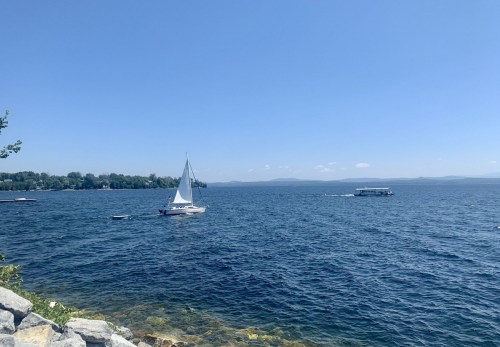 A sailboat glides across a calm blue lake under a clear sky, with distant hills visible on the horizon.