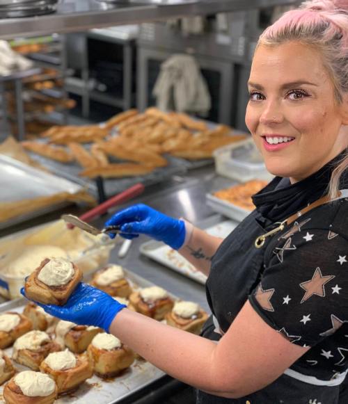 A smiling baker in gloves decorates cinnamon rolls with frosting in a busy kitchen filled with baked goods.
