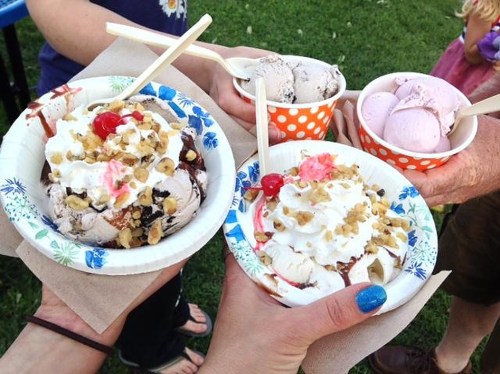 Two hands hold ice cream sundaes topped with whipped cream, cherries, and nuts, alongside cups of ice cream.