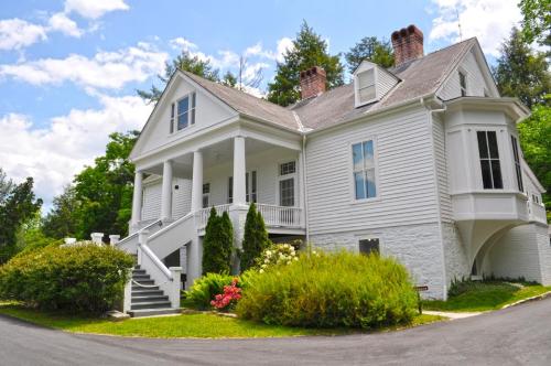 A large white house with a porch, surrounded by greenery and colorful flowers under a partly cloudy sky.