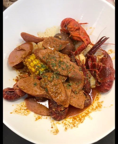 A bowl of seafood featuring crawfish, sausage, corn, and a rich, spicy sauce, garnished with herbs.