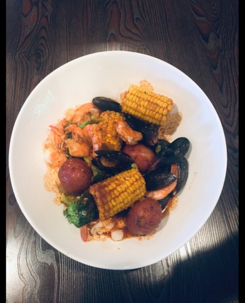 A bowl of seafood featuring shrimp, mussels, corn, potatoes, and broccoli on a wooden table.