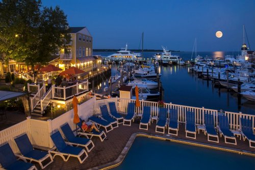 A marina at night with boats, a pool, and a full moon reflecting on the water, surrounded by lit buildings and trees.
