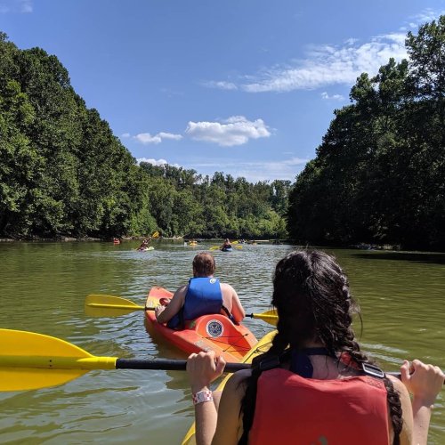 Two people in kayaks paddle down a calm river surrounded by lush green trees under a clear blue sky.