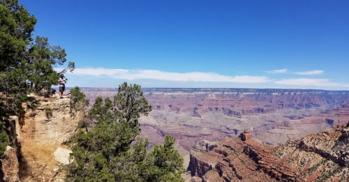A person stands on a rocky ledge overlooking the vast Grand Canyon under a clear blue sky.