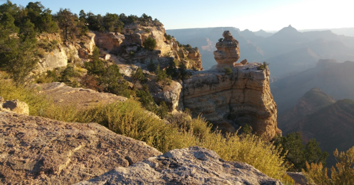A scenic view of rugged cliffs and rock formations at sunrise, with greenery in the foreground and mountains in the distance.