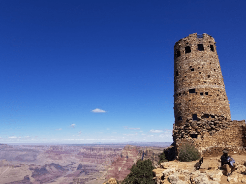 A stone tower stands on the edge of the Grand Canyon, with a clear blue sky and colorful canyon layers in the background.