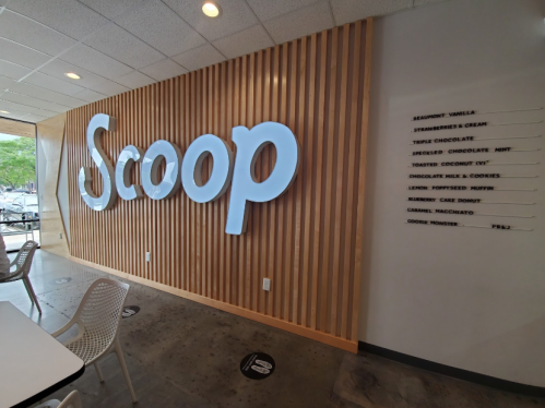 Interior of an ice cream shop featuring a wooden wall with the word "Scoop" and a menu of flavors on the side.