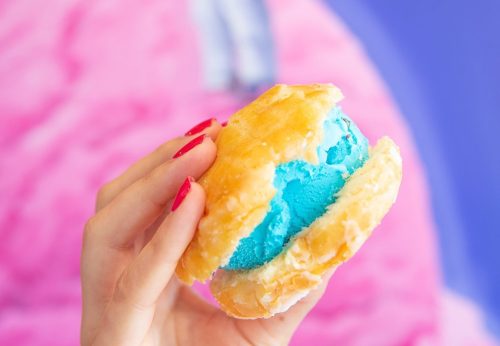 A hand holding a colorful ice cream sandwich with bright blue ice cream between two golden, flaky pastries.