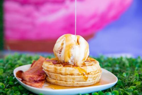 A waffle topped with a scoop of ice cream and drizzled with syrup, served with crispy bacon on the side.
