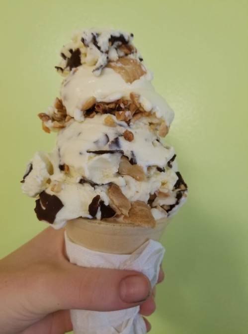 A hand holding a tall ice cream cone topped with layers of vanilla ice cream, chocolate drizzle, and crunchy toppings.
