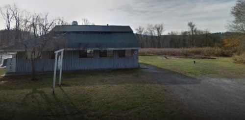 A weathered building with a gray roof, surrounded by trees and grass, set against a cloudy sky.