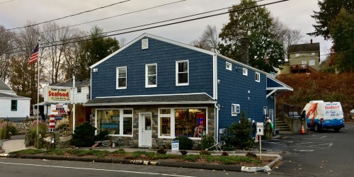 A blue seafood market building with a sign, parked delivery van, and trees in the background on a cloudy day.