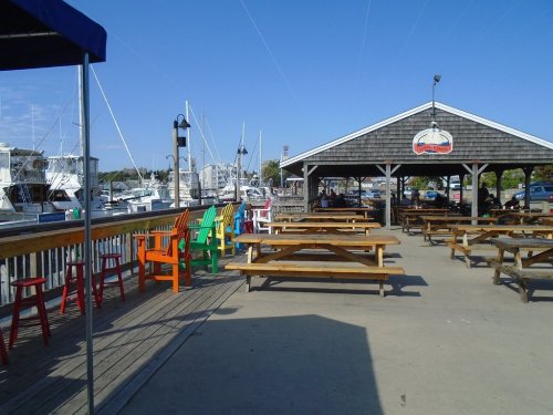 Colorful chairs line a wooden deck by a marina, with boats docked and a covered seating area in the background.