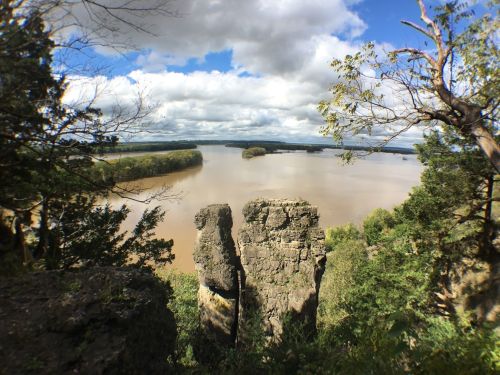 A scenic view of a river surrounded by greenery and rocky formations under a partly cloudy sky.