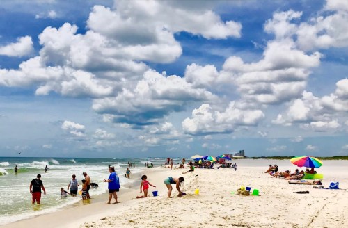 A sunny beach scene with people playing in the sand and water, colorful umbrellas, and fluffy clouds in the sky.