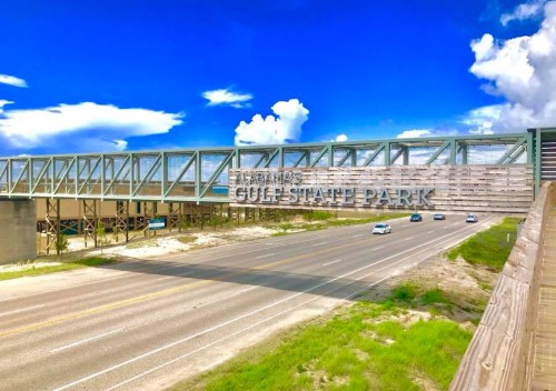 A bridge over a highway leading to Gulf State Park, with blue skies and scattered clouds in the background.