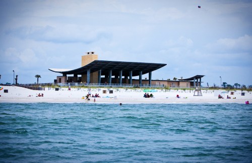 A modern beach facility with a large roof, surrounded by sandy shores and people enjoying the water and sun.