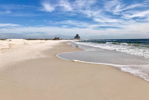 A serene beach scene with gentle waves, soft sand, and a distant skyline under a blue sky with wispy clouds.