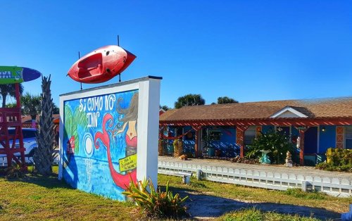 Colorful mural on a sign outside a beach-themed building, with a red kayak perched on top against a clear blue sky.