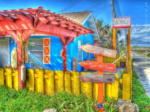 Colorful beachside shop with a red pergola, yellow fence, and directional signs for local attractions. "Vacancy" sign visible.
