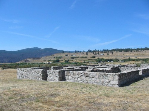 Ruins of stone structures in a grassy landscape, with mountains and a clear blue sky in the background.