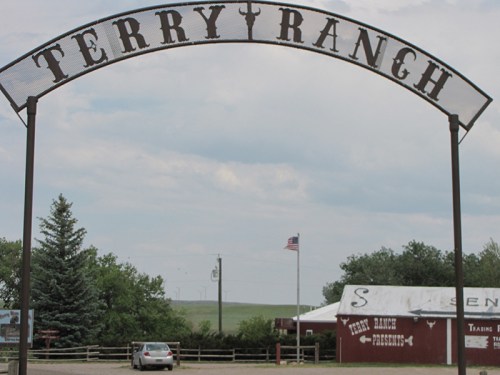 Sign for Terry Ranch with a cloudy sky, trees, and a building in the background. A car is parked nearby.