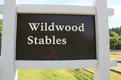 Sign for "Wildwood Stables" displayed on a white wooden post against a green landscape.