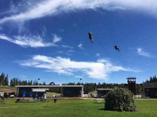 Two people ziplining against a blue sky with clouds, with a grassy area and buildings in the background.