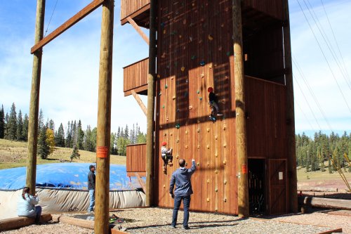 Two children climb a wooden rock wall while an adult supervises. A blue inflatable mat is nearby, surrounded by trees.