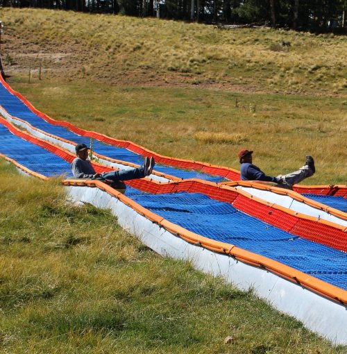 Two people relax on a colorful, sloped water slide in a grassy field surrounded by trees.