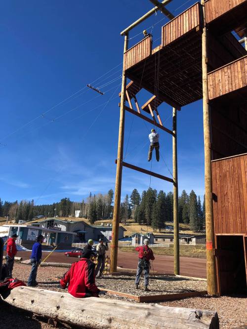 A person rappels down a wooden tower while others observe and assist on the ground in a training environment.