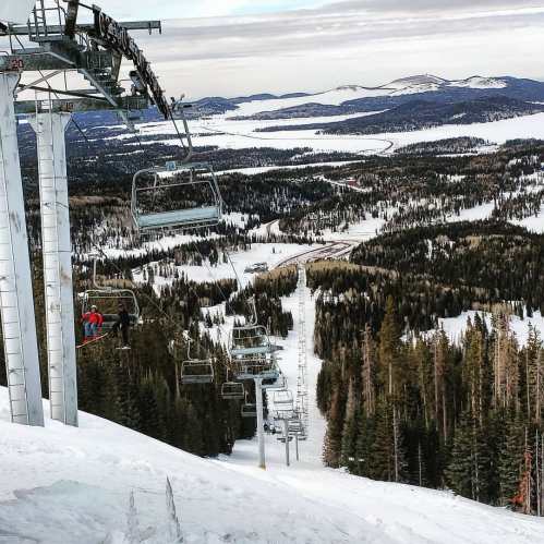 A ski lift ascends a snowy mountain, surrounded by evergreen trees and a vast winter landscape.