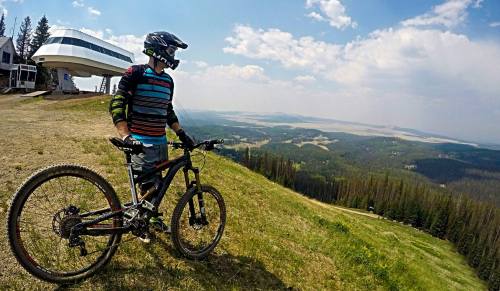 A mountain biker in gear stands on a hillside, overlooking a vast landscape with trees and distant mountains.