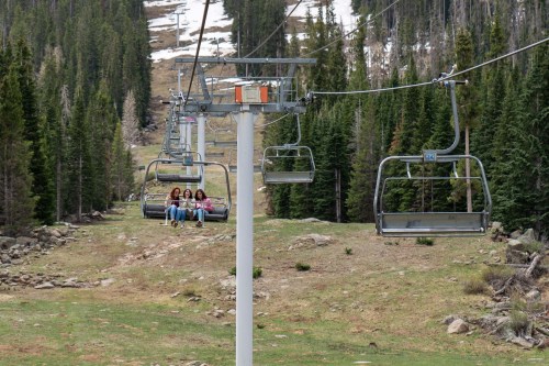 Three people sit on a ski lift, surrounded by trees and a grassy landscape, with patches of snow in the background.