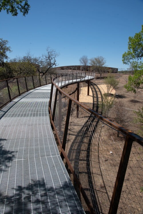 A winding metal walkway curves through a dry landscape under a clear blue sky, surrounded by sparse trees.
