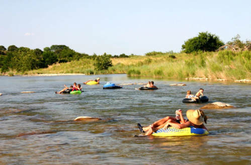 People relaxing on inner tubes in a river, surrounded by greenery and a clear blue sky.