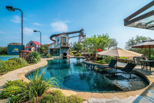 A scenic pool area featuring a waterslide, lush greenery, and lounge chairs under umbrellas on a sunny day.