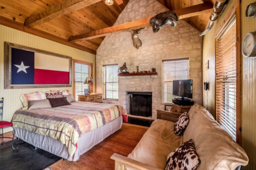 Cozy bedroom with a Texas flag, stone fireplace, wooden beams, and rustic decor, featuring a bed and a sofa.