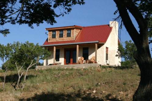 A charming house with a red metal roof, set on a grassy hill surrounded by trees under a clear blue sky.