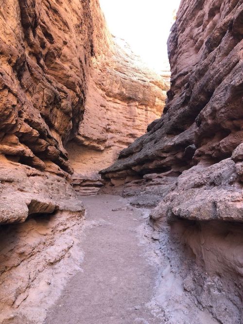 A narrow canyon with steep, rocky walls and a sandy path leading through it, illuminated by sunlight.
