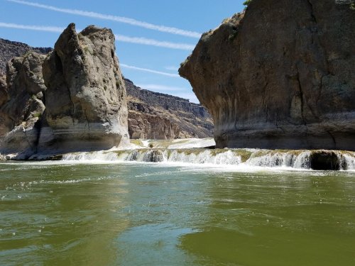 A serene river flows between large rock formations under a clear blue sky with wispy clouds.