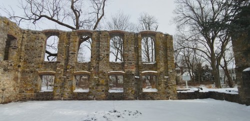 Snow-covered ruins of a stone building with arched windows, surrounded by bare trees in a winter landscape.