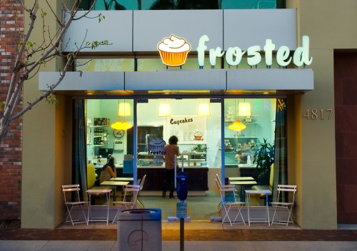 A storefront of "Frosted" with a cupcake sign, featuring outdoor seating and a cozy interior.