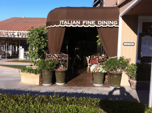 Entrance of an Italian fine dining restaurant with potted plants and a brown awning, next to a Chinese food place.