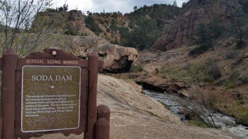 A scenic view of Soda Dam with a sign detailing its geological features and a flowing river in the background.