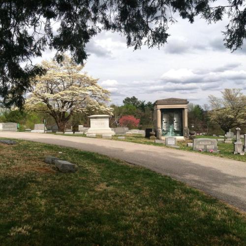 A peaceful cemetery scene with gravestones, a mausoleum, and blooming trees under a cloudy sky.